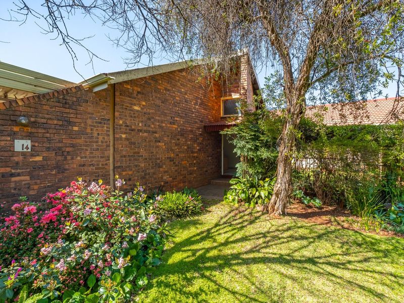 Front exterior view of a brick townhouse featuring a landscaped garden, mature trees, and a covered entrance area.