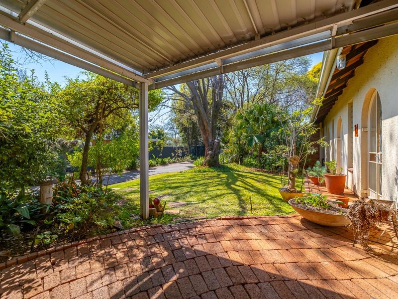 A covered brick-paved patio area overlooking a lush, established garden with mature trees and arched access to the main dwelling.