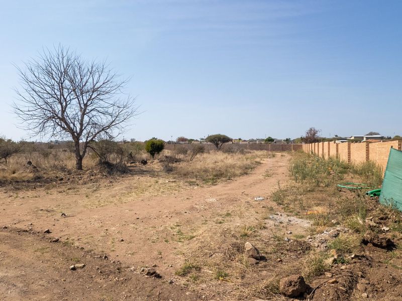 A view of a vacant commercial land plot featuring a dirt access path and a perimeter brick boundary wall.