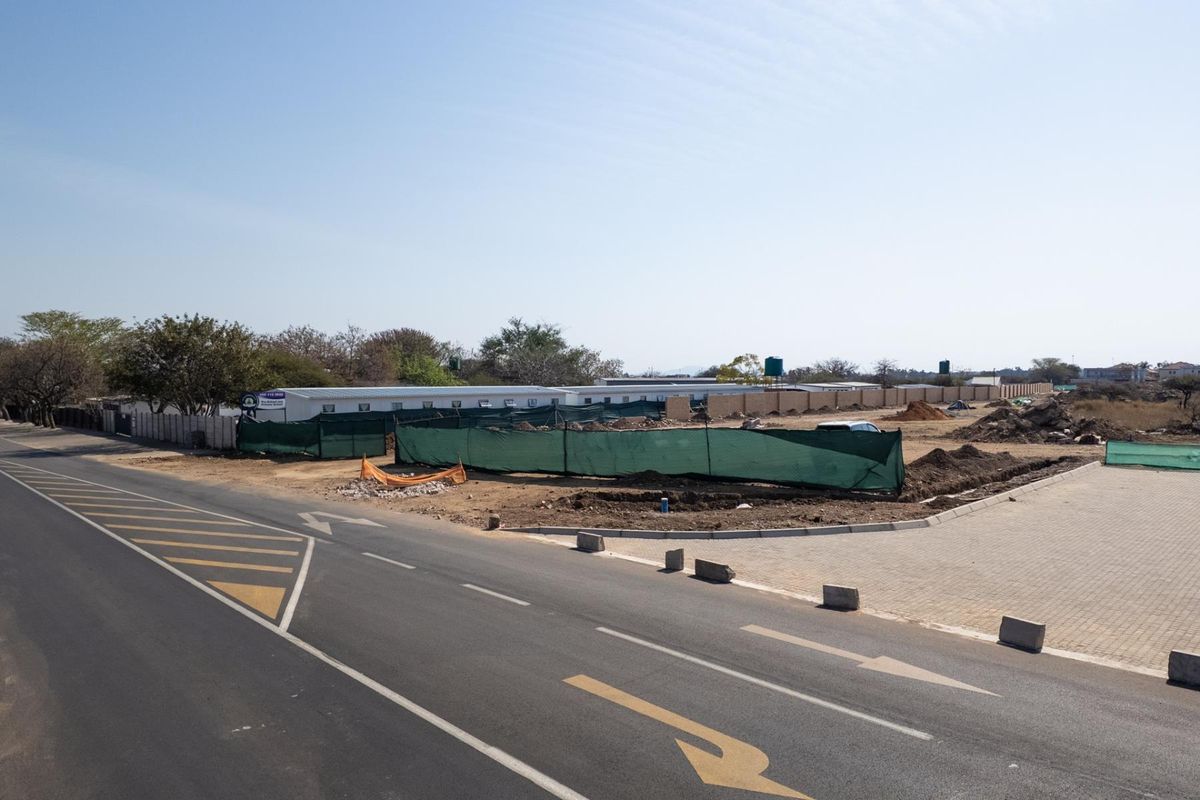 A street-level view of a vacant commercial land plot featuring perimeter walling and temporary construction fencing along a paved road.