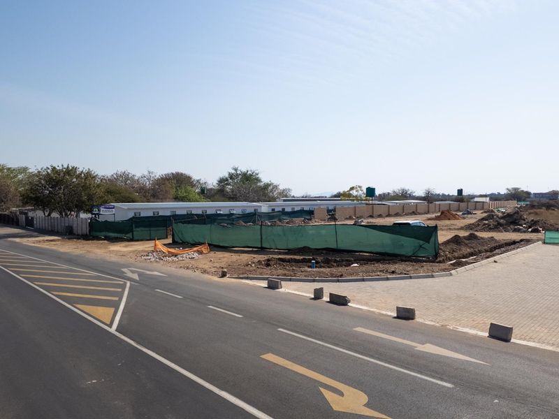 A street-level view of a vacant commercial land plot featuring perimeter walling and temporary construction fencing along a paved road.