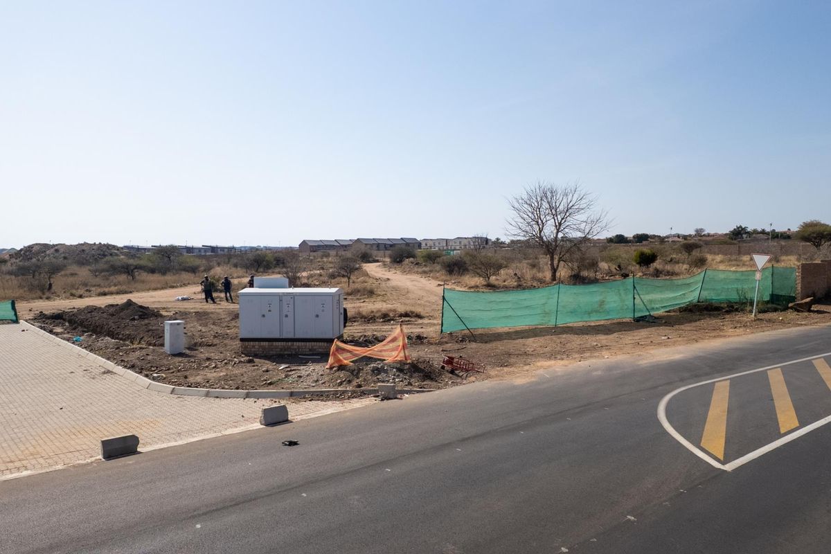 A view of a vacant commercial land plot featuring a paved driveway entrance, electrical infrastructure, and perimeter fencing.