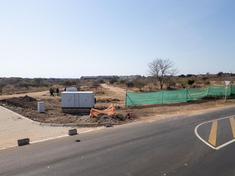 A view of a vacant commercial land plot featuring a paved driveway entrance, electrical infrastructure, and perimeter fencing.