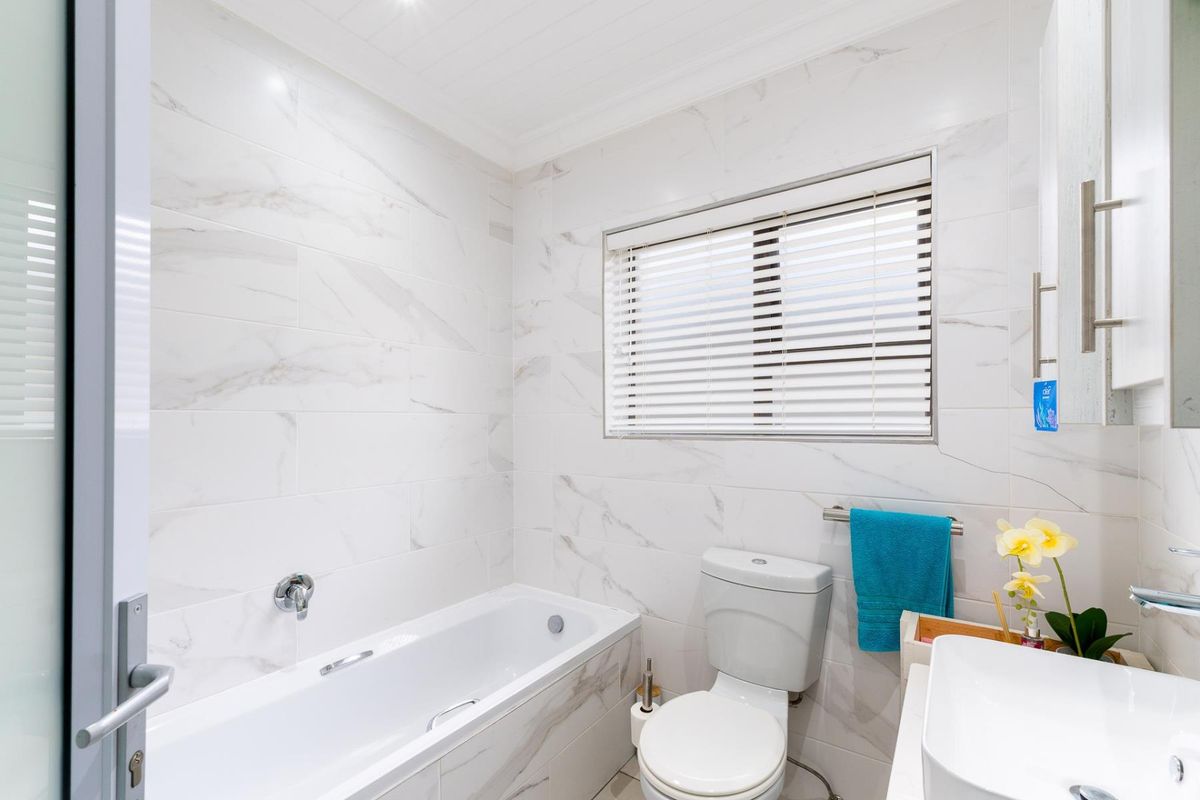 Modern bathroom featuring white marble-look wall tiles, a built-in bathtub, a wall-mounted faucet, a toilet, and a vanity basin with venetian blinds.