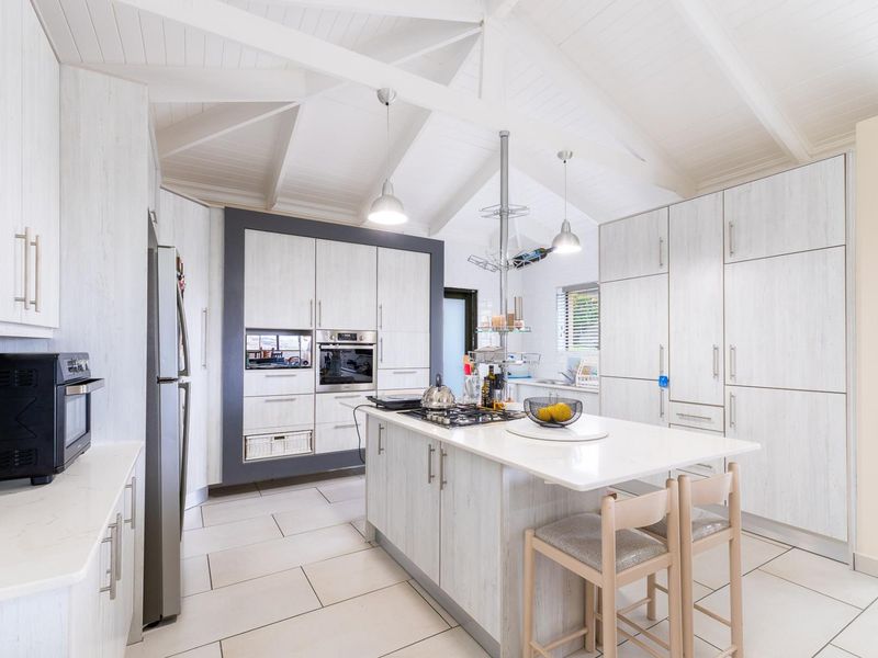 Modern kitchen featuring a central island with gas hob, built-in appliances, light wood-grain cabinetry, and a high vaulted ceiling with exposed beams.