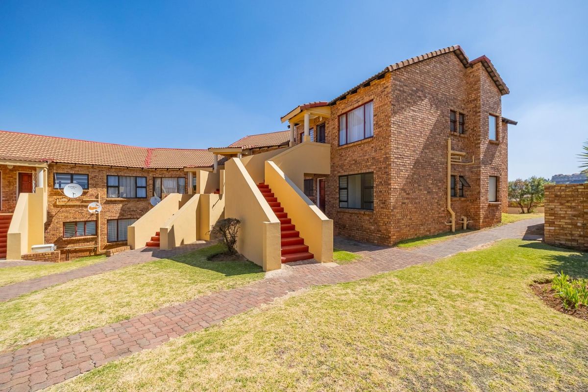Rear view of a face brick townhouse complex featuring external staircases, paved walkways, and established lawn areas under a clear blue sky.