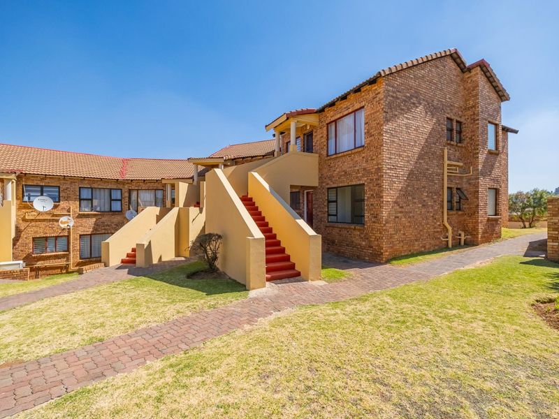 Rear view of a face brick townhouse complex featuring external staircases, paved walkways, and established lawn areas under a clear blue sky.