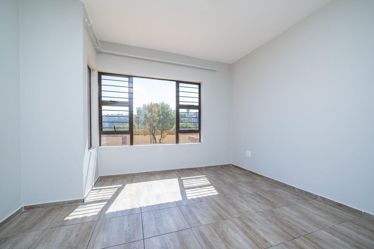 A bright, empty bedroom featuring neutral tiled flooring and large aluminium-framed louvered windows providing natural light.