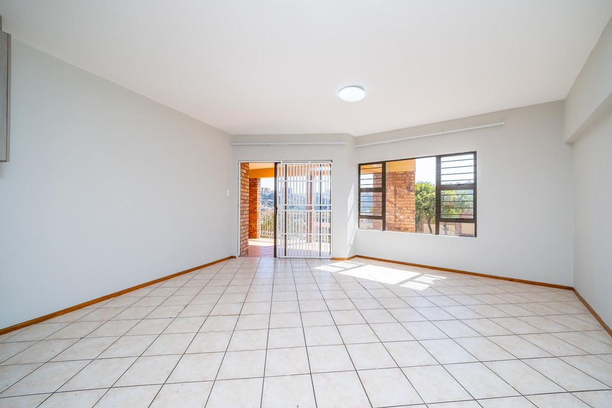 A bright, empty living room featuring neutral tiled flooring, large windows, and a sliding door leading to an outdoor patio area.