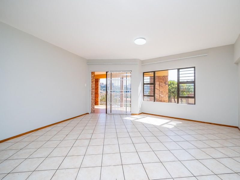 A bright, empty living room featuring neutral tiled flooring, large windows, and a sliding door leading to an outdoor patio area.