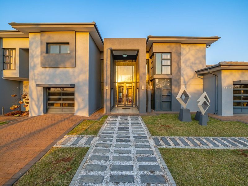 A modern double-storey residence featuring a striking glass entrance, paved driveway, and a well-maintained front garden with a stone walkway.