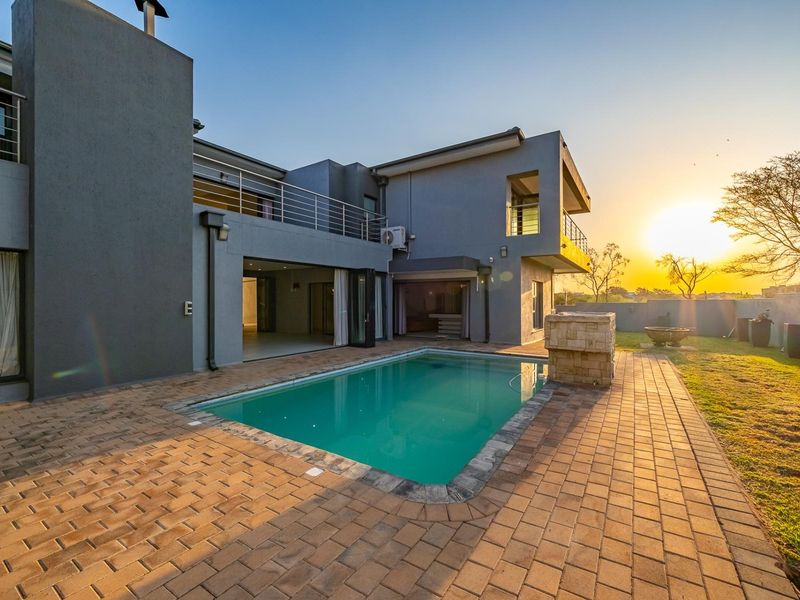 A modern double-story home exterior featuring a paved pool area, stone feature wall, and an upper-level balcony, captured during sunset.