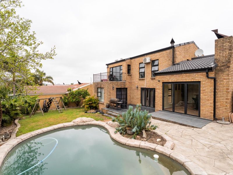 Rear view of a multi-story brick house featuring a swimming pool, paved patio area, upper-level balcony, and established garden.
