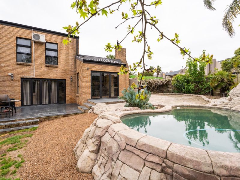 Rear exterior view featuring a brick facade, tiled patio area, and a rock-feature swimming pool set within a landscaped garden.