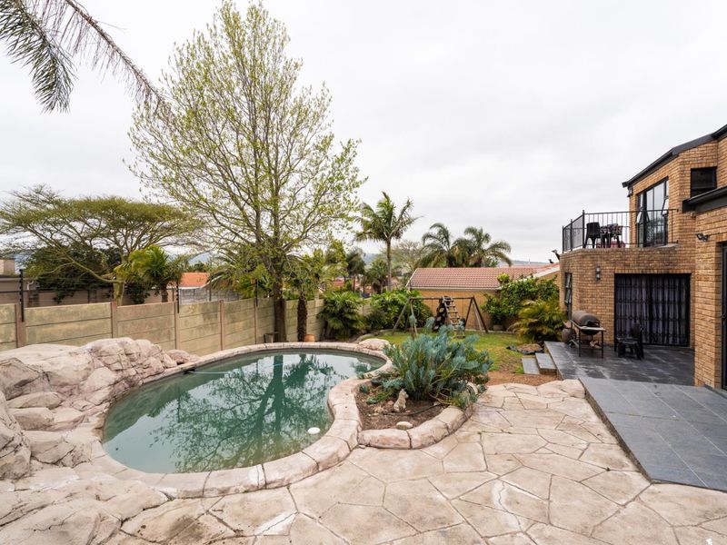 Rear garden featuring a rock-edged swimming pool, paved patio area, and a brick-built house with an upper-level balcony.