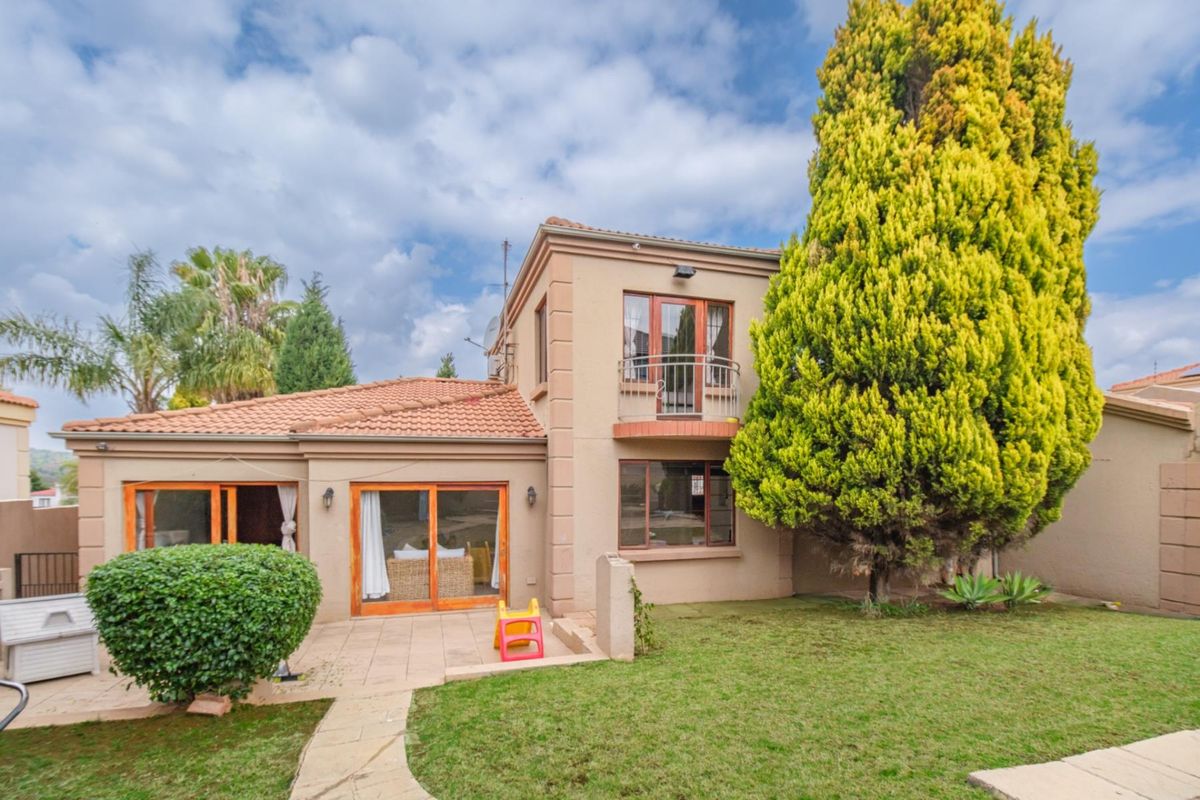 Rear view of a two-story townhouse featuring a paved patio, sliding glass doors, a small upper-level balcony, and a well-maintained garden area.