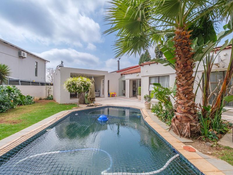 A well-maintained outdoor pool area featuring a swimming pool with a safety net, adjacent covered patio, and established garden landscaping.