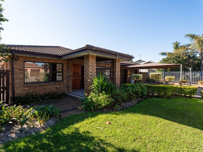 A well-maintained face brick house featuring a tiled roof, paved entrance walkway, and a convenient carport, set within a neat garden with perimeter fencing.