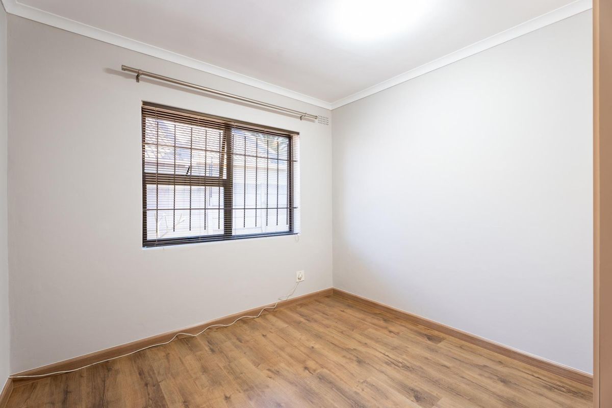 A bright, empty bedroom featuring laminate wood flooring, neutral walls, and a window fitted with security bars and a curtain rod.