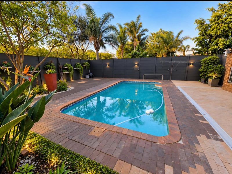 A well-maintained outdoor pool area featuring brick paving, perimeter fencing, and integrated wall lighting, surrounded by landscaped garden elements.