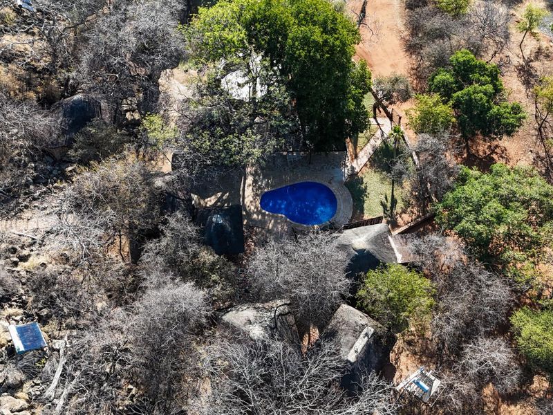 Aerial view of a farm property featuring a swimming pool, thatched roof buildings, and natural landscape with rock formations and trees.