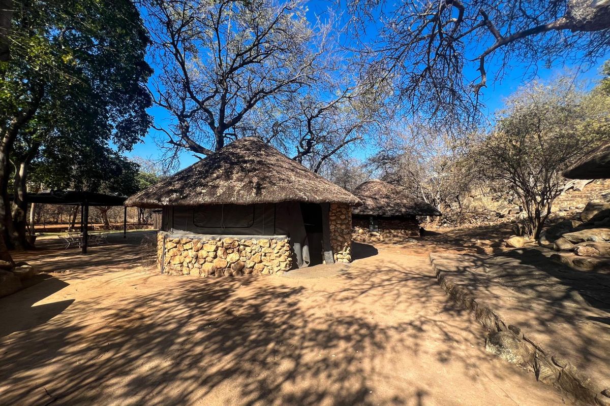 A rustic farm-style dwelling featuring traditional thatched roofing and stone masonry walls, set within a natural, wooded outdoor environment.
