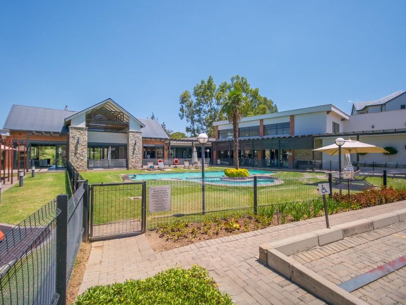 A well-maintained communal pool area featuring a landscaped garden, stone-clad building architecture, and paved walkways within a secure residential complex.