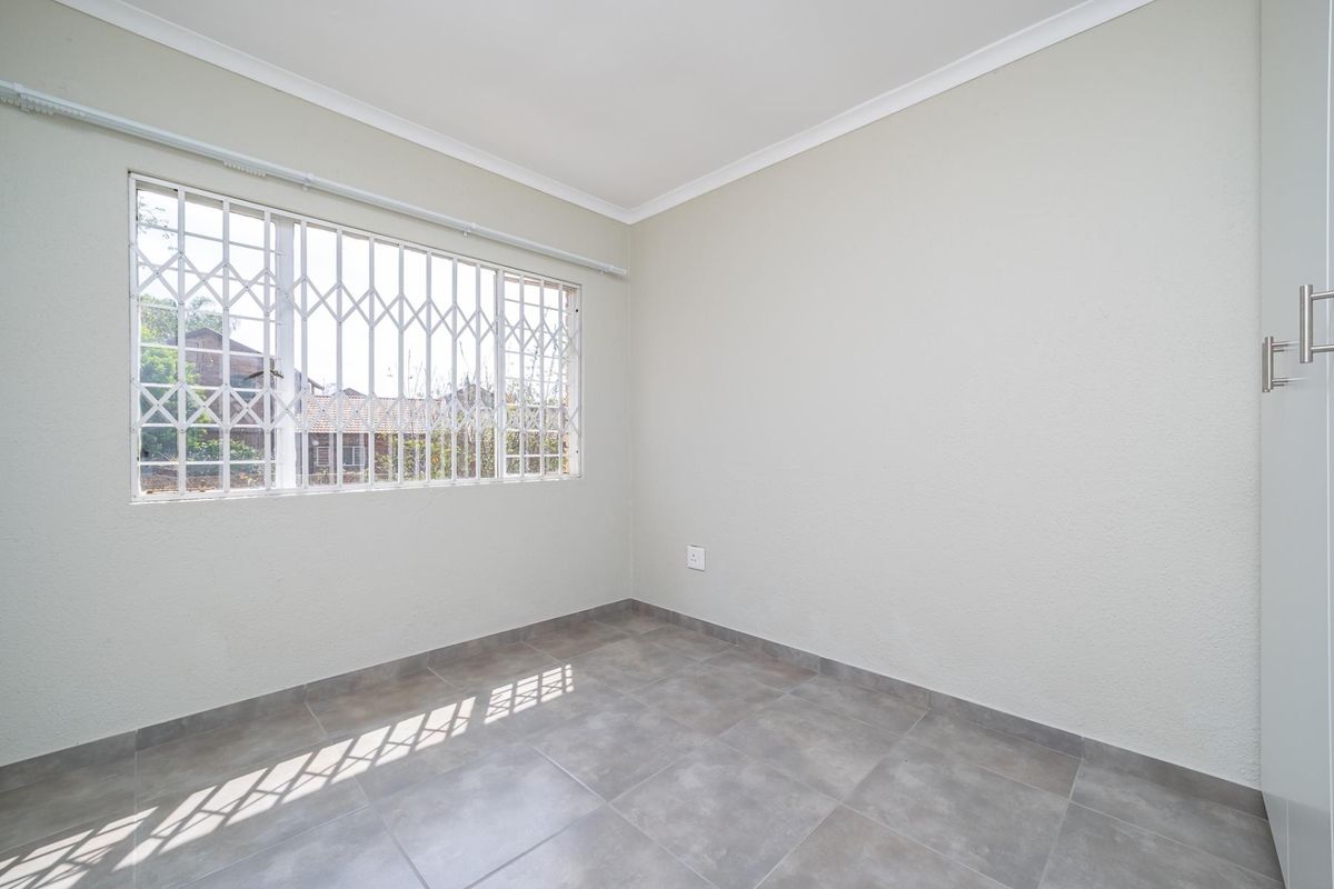 A bright, empty bedroom featuring neutral tiled flooring, a built-in cupboard, and a window fitted with a security gate.