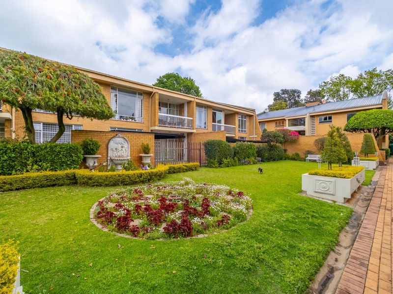 A well-maintained rear garden area featuring landscaped lawns, flower beds, a decorative fountain, and paved walkways leading to apartment building balconies.