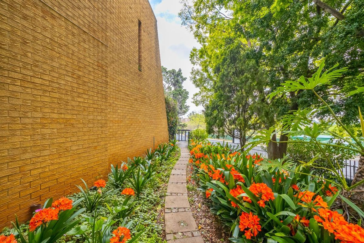 A well-maintained exterior side path featuring a paved walkway bordered by vibrant orange clivia flowers and mature trees alongside the building's brick facade.