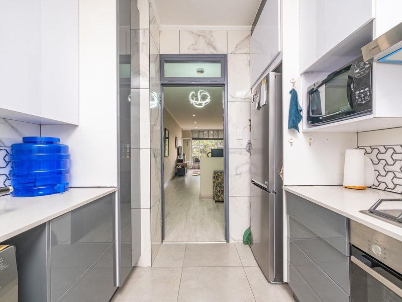 Modern galley-style kitchen featuring high-gloss cabinetry, stone countertops, integrated appliances, and stylish marble-look wall tiling, leading through to the living area.