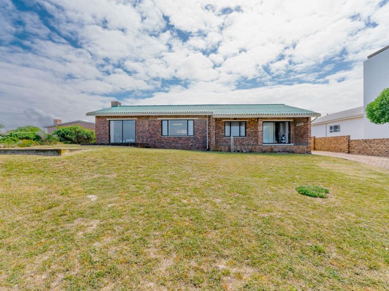 A rear view of a single-story face brick house featuring a green corrugated metal roof, large windows, and a spacious lawn area.