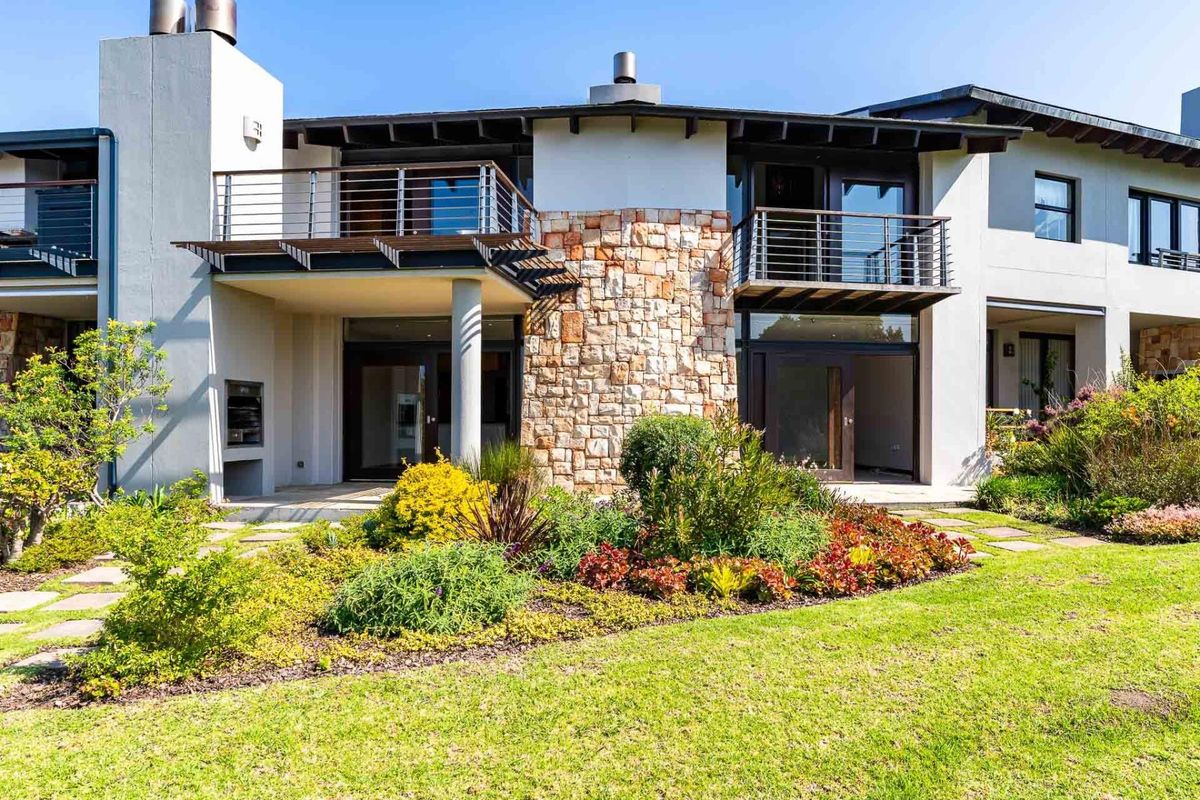 Rear elevation of the apartment featuring a landscaped garden, paved patio with built-in braai, and upper-level balconies with stainless steel balustrades.