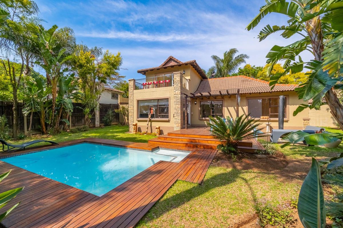 Rear view of the house featuring a swimming pool, timber decking, landscaped garden, and an upper-level balcony with stone-clad pillars.