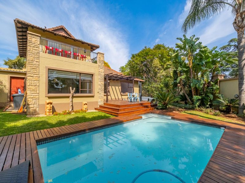 Rear exterior view featuring a swimming pool with timber decking, a pergola-covered patio, and an upper-level balcony with stone-clad architectural detailing.