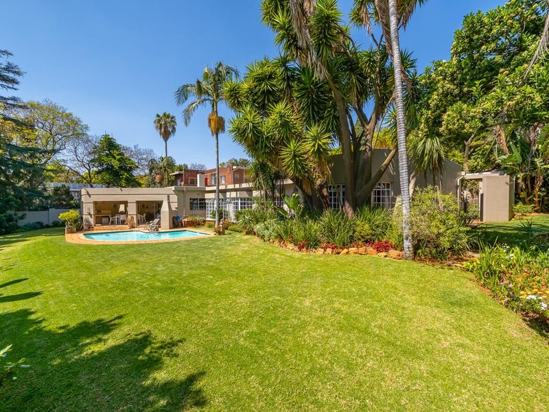Spacious rear garden featuring a well-maintained lawn, mature trees, a swimming pool with paved surround, and a covered patio area.