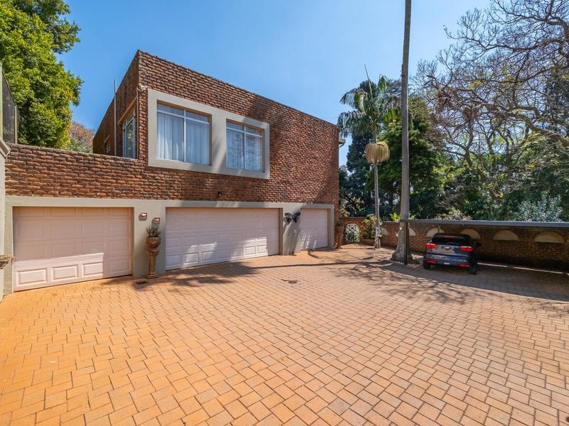Front exterior view featuring a triple garage, paved driveway, and additional carport space, set against a brick facade and mature trees.
