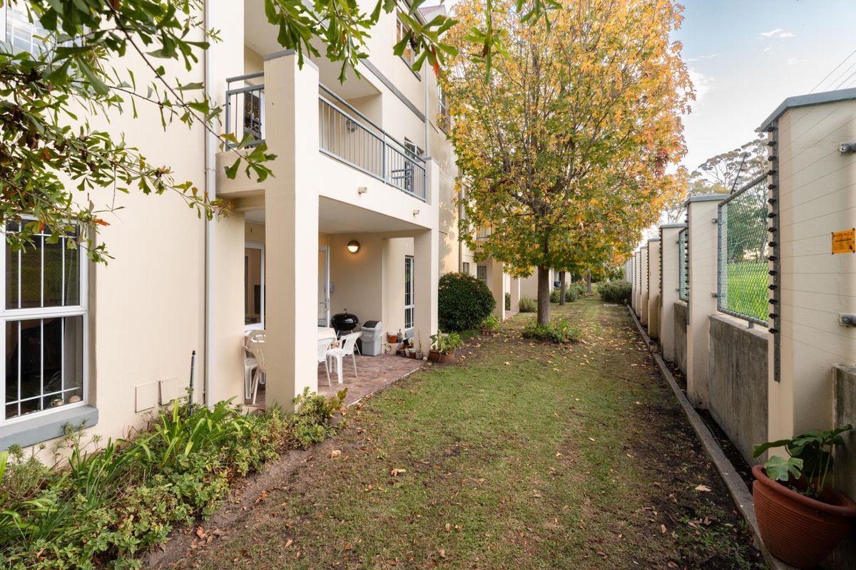 Rear view of the apartment building featuring a ground-floor paved patio, upper-level balcony with metal railings, and a landscaped garden area with perimeter security fencing.