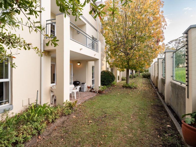 Rear view of the apartment building featuring a ground-floor paved patio, upper-level balcony with metal railings, and a landscaped garden area with perimeter security fencing.