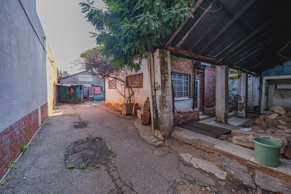 Rear exterior area featuring a paved driveway, brick building structure, and a covered patio space with visible signs of wear and structural deterioration.
