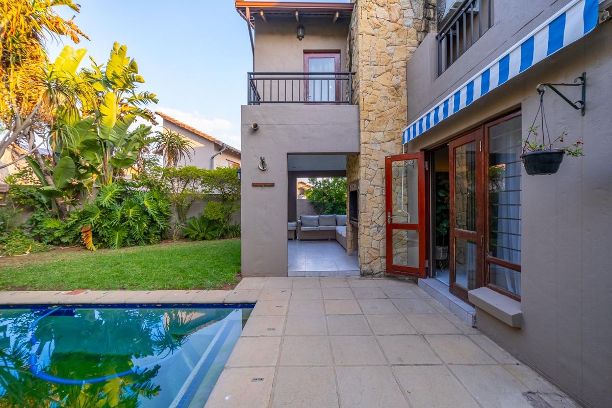 Rear exterior view featuring a swimming pool, paved patio area, covered outdoor entertainment space with built-in braai, and stone-clad architectural detailing.