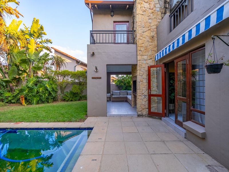 Rear exterior view featuring a swimming pool, paved patio area, covered outdoor entertainment space with built-in braai, and stone-clad architectural detailing.