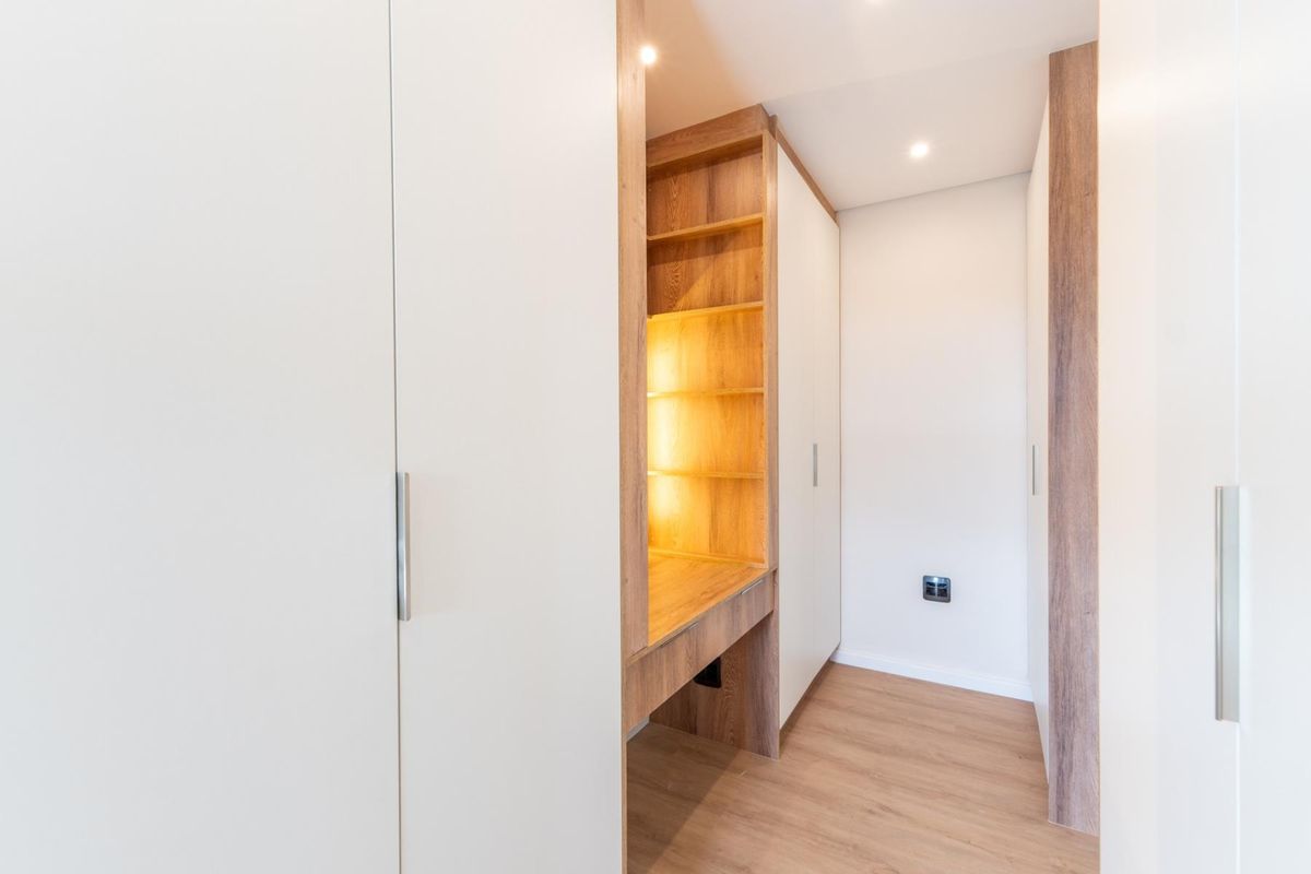 A modern, empty bedroom featuring extensive built-in white cabinetry, integrated wooden shelving with warm accent lighting, and light-toned laminate flooring.