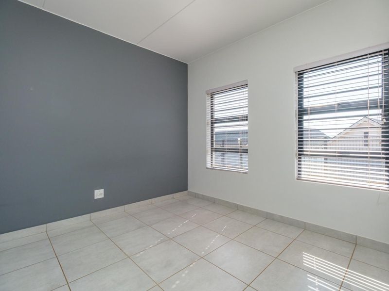 A bright, empty bedroom featuring neutral tiled flooring, an accent wall, and dual windows fitted with venetian blinds.