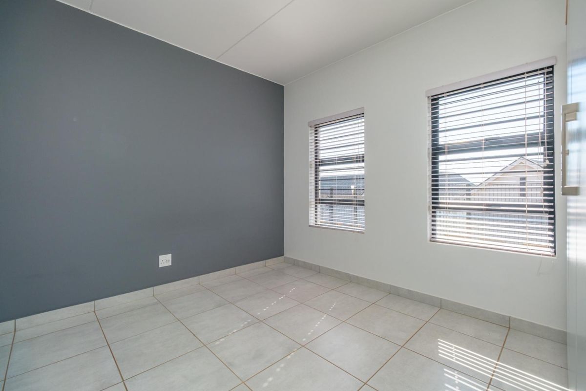 A bright, empty bedroom featuring neutral tiled flooring, an accent wall, and dual windows fitted with venetian blinds.