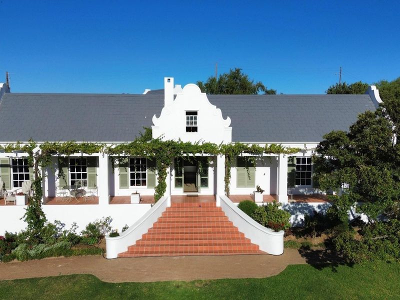 An aerial view of a well-maintained Cape Dutch style home featuring a prominent central gable, tiled entrance staircase, and a spacious covered veranda.