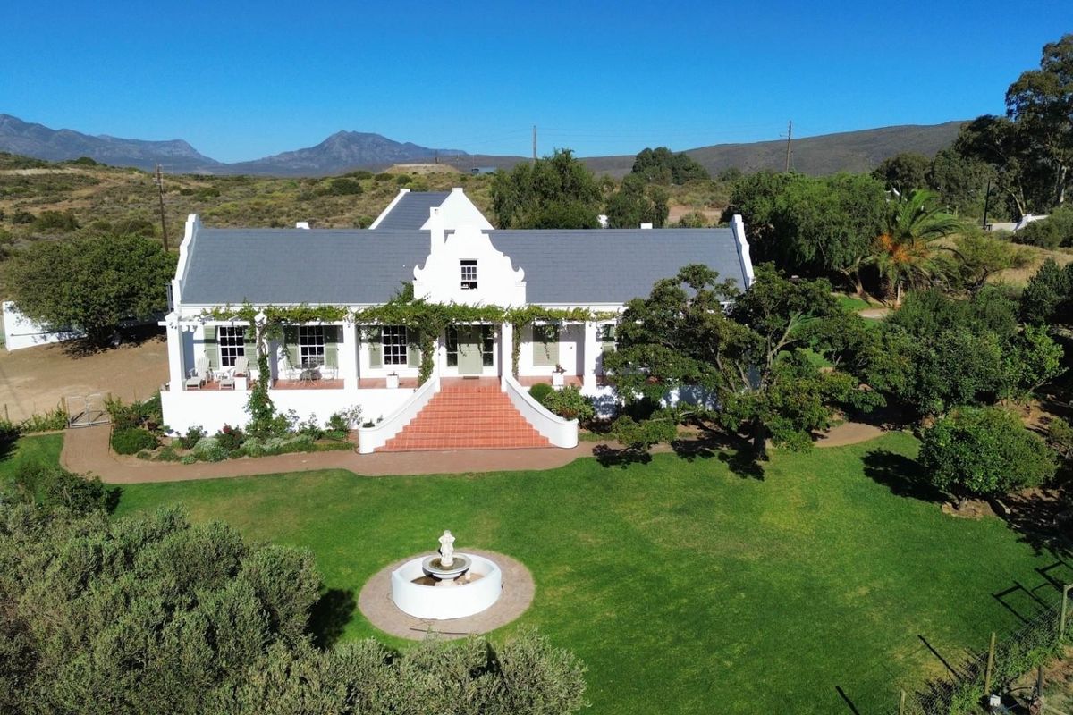 Aerial view of a classic Cape Dutch style house featuring a prominent gable, terracotta entrance stairs, covered veranda, and a well-maintained garden with fountain.