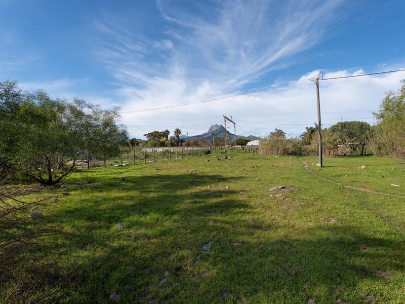 A vacant residential plot featuring grassy terrain, perimeter wire fencing, and utility infrastructure with mountain views in the background.