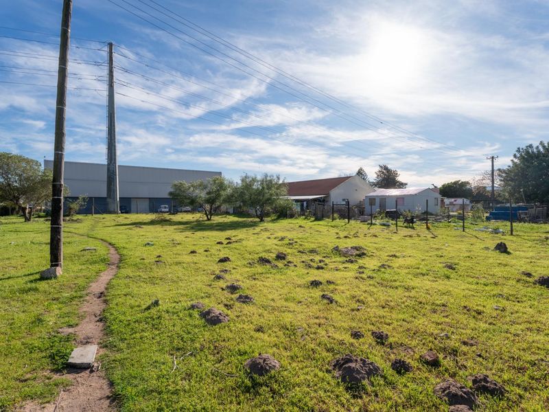 A view of a vacant residential plot featuring uneven grassy terrain, wire fencing, and overhead utility lines with neighboring structures in the background.