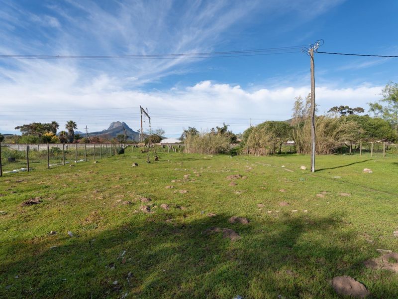 A vacant residential plot featuring open grassy terrain, perimeter wire fencing, and existing utility infrastructure with mountain views in the distance.